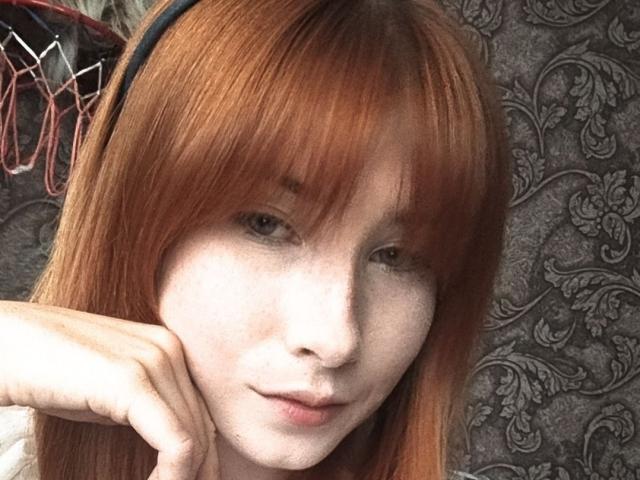 KasandraGrothe has red hair, freckles, and a slight smile with her chin resting on her hand. She's wearing a black headband with red strings.