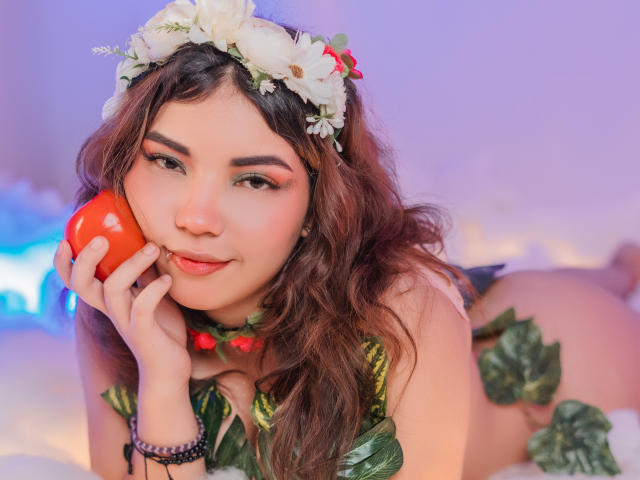 SweetyAliceHotX with curly brown hair, floral headband, and green leaf dress, holding a red apple, smiling, wearing a bead necklace and bracelet, lying on a bed.