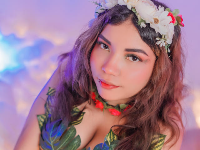 SweetyAliceHotX with brown wavy hair, wearing a flower headband, green leafy dress, red flower necklace, and red lipstick, showing her tits, looking cute and sexy.