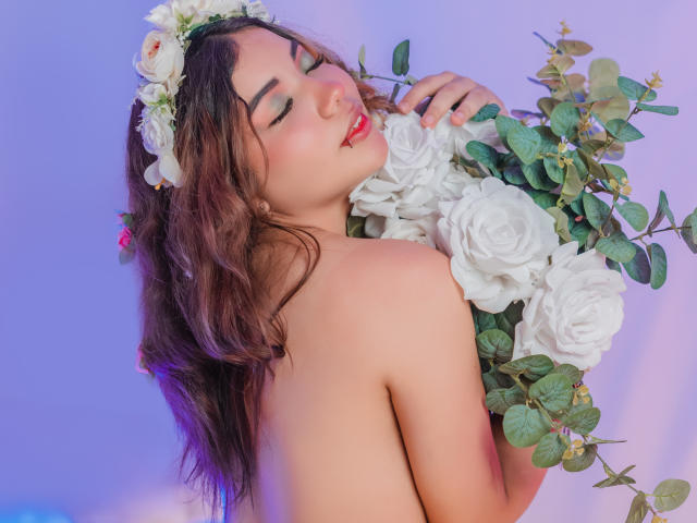 SweetyAliceHotX with wavy brown hair, floral headpiece, red lipstick, holding white roses and eucalyptus, topless, smiling, eyes closed, soft purple and blue lighting.