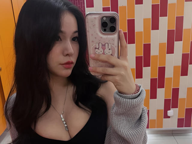 ShelliQuirin with long black hair, red lips, black top showing big tits, gray sweater, holding phone with bunny case, taking mirror selfie, orange and red wall in background.