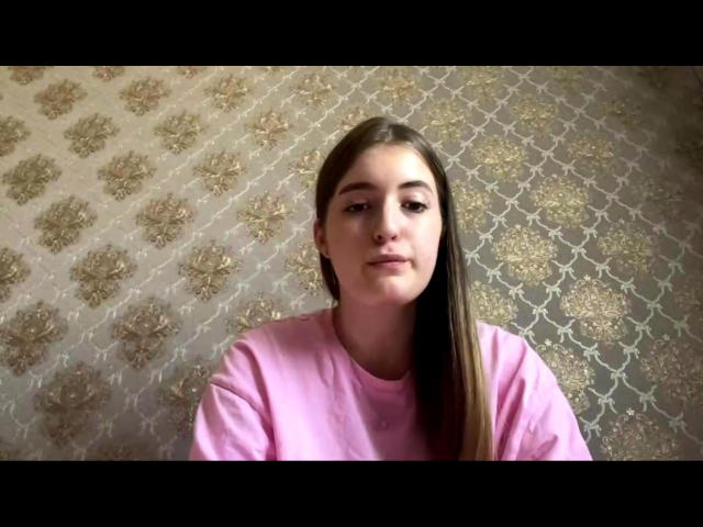 GabrielKahoun has long brown hair, a pink shirt, and a neutral expression, standing against a patterned wall. Her tits are small, and she's looking straight at the camera.