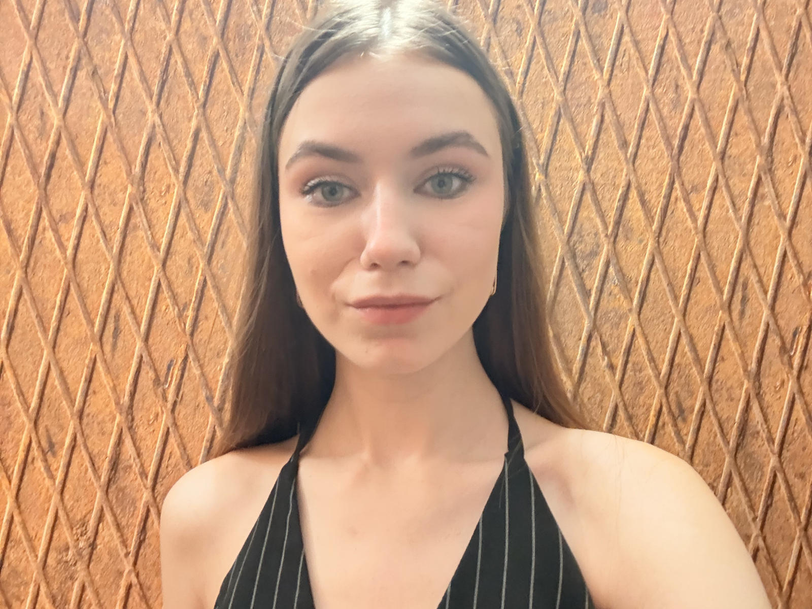CaritaShover with long brown hair, green eyes, and pouty lips, wearing a black pinstripe halter top, looking directly at the camera with a slight smile.