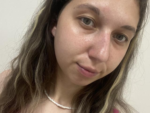 Sanella's face with light brown wavy hair, small nose, slightly open mouth, and a white beaded necklace. She has a gentle smile and a slightly shiny, smooth complexion.