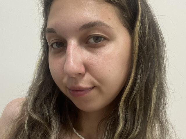 Sanella has light brown skin, wavy dark brown hair with blonde highlights, brown eyes, and a slight smile. She's wearing a white necklace. Her face is centered.