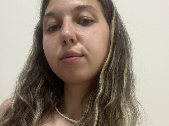Sanella has wavy brown hair with blonde highlights, a light skin tone, and is wearing a pearl necklace. Her small tits are partially visible. She's smiling slightly.