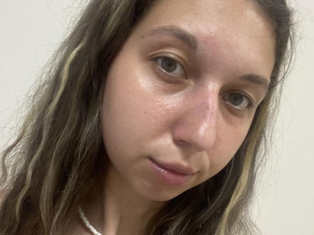 Sanella has light brown, wavy hair with blonde highlights, a slightly shiny, pinkish-tan face, and a subtle, closed-lip smile. Her blue eyes are slightly squinted.