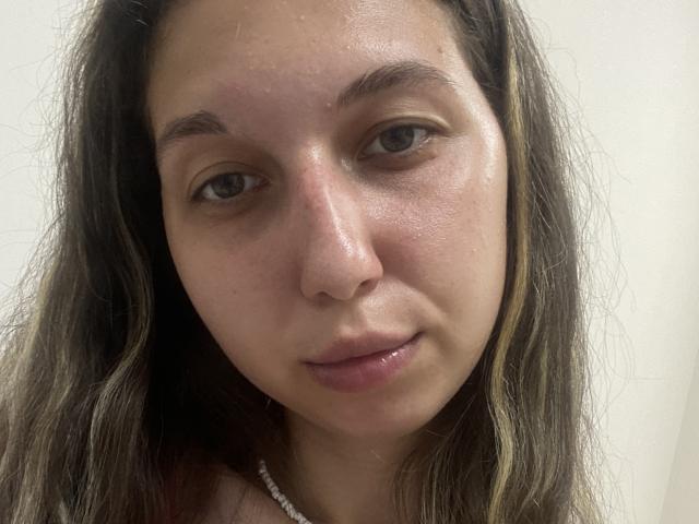 Sanella's face, showing a slight smile with glossy pink lips, has light brown skin, dark brown wavy hair with blonde highlights, and small eyebrows. She wears a white necklace.