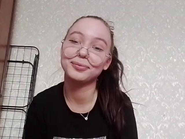 FayStorbeck with glasses, black shirt, and ponytail, sticking out tongue, showing cute face, white wall, metal rack on left, soft smile, and slightly messy hair.