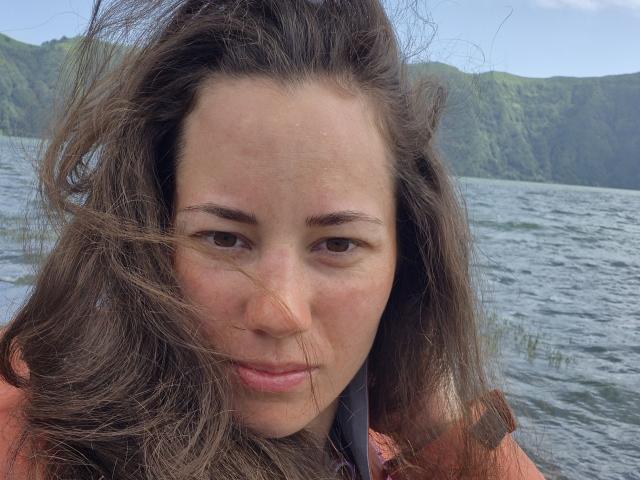 ArianeEsther a les cheveux bruns ondulés qui flottent dans le vent, des yeux bruns et un sourire discret, elle porte une veste orange, avec un lac et des collines verdoyantes en arrière-plan..