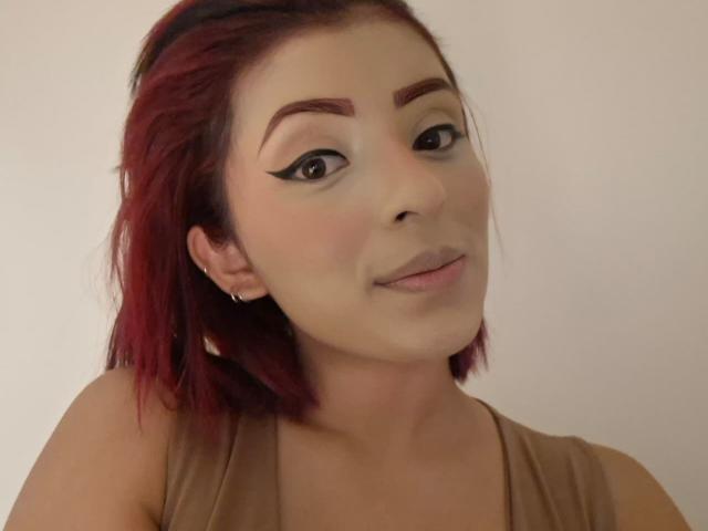 LizBonnet met rood haar, donkere oogschaduw, en subtiele make-up, draagt een beige top, kleine oorringen en heeft een lichte glimlach, kijkend iets naar rechts..