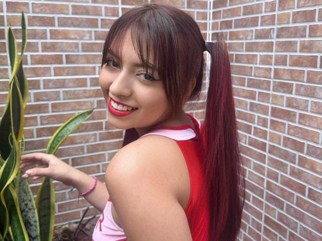 BrendaaPierce with red pigtails, smiling, wearing a red sleeveless top, showing her shoulder, standing next to a green plant, brick wall in background, wearing pink bracelet.