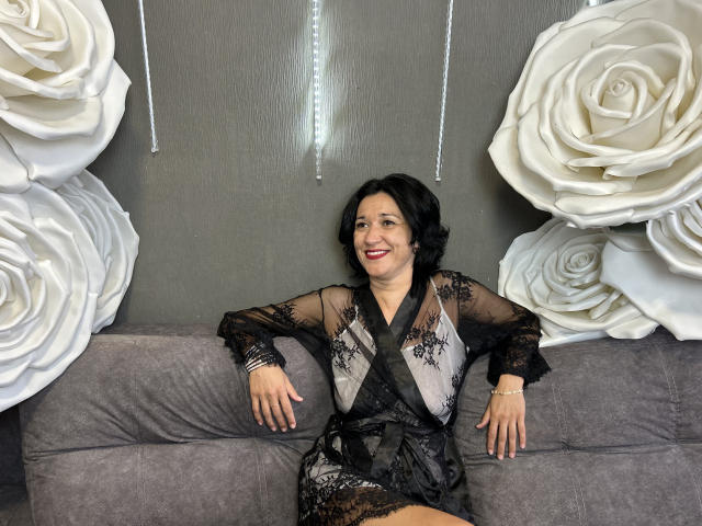 CameronEnns is sitting on a gray sofa, wearing a black lace robe, showing off her tits, with white rose decorations on both sides. She's smiling with red lipstick.
