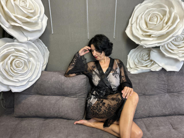 CameronEnns sits on a grey couch, wearing a black lace robe, showing her tits and ass, with her short black hair, hand on her head, and leg raised.