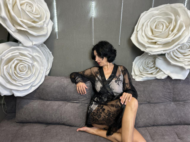 CameronEnns, short black hair, wearing black lace robe, sitting on gray couch, legs crossed, white rose decorations on wall, looking down with slight smile, medium tits, fair skin.