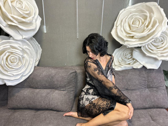 CameronEnns, with short black hair, sits on a gray sofa, wearing a black lace dress, showing her ass, in a room with large white rose decorations.