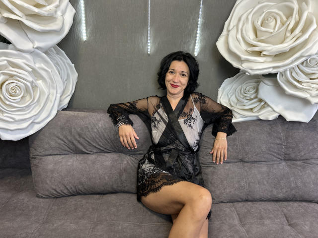 CameronEnns with short black hair, wearing a sheer black lace dress, sitting on a gray sofa, legs crossed, white rose wall decor on both sides, smiling.
