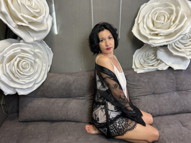 CameronEnns, a short-haired black woman, kneels on a gray couch, wearing a white tank top and black lace robe, exposing her white lace panties and her ass.