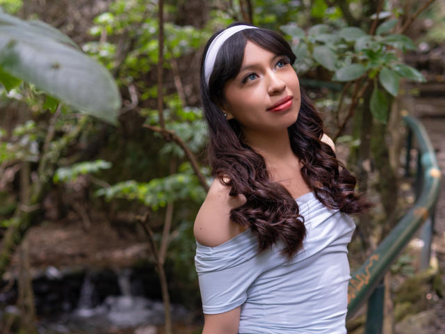 ZoeRedds with curly brown hair, white headband, off-shoulder grey top, looking up with a slight smile, standing in a forest with a small waterfall in the background.