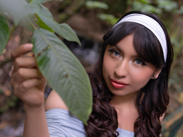 ZoeRedds with dark wavy hair, white headband, blue eyes, holding a green leaf, wearing a gray top, soft pink lips, small earrings, looking at camera with gentle smile.