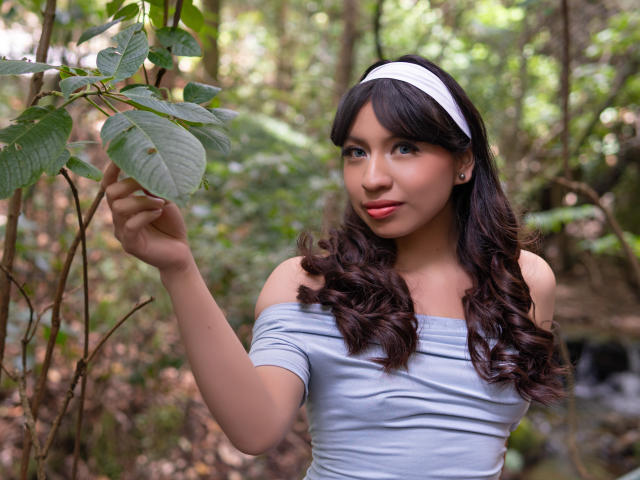 ZoeRedds with curly brown hair, white headband, and off-shoulder gray top, touching green leaves with her right hand, has red lipstick and small earrings, standing in a forest.