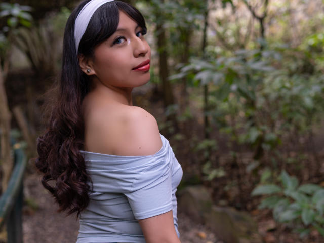 ZoeRedds with long, wavy black hair, wearing a light blue off-shoulder top and white headband, looking over her shoulder with red lips and small earrings. Her tits are slightly visible.