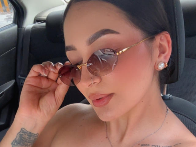 PamelaLiss with tan skin, dark eyebrows, round pink-tinted sunglasses, small nose ring, pearl earrings, and tattoos on her wrist, sitting in a car, holding her sunglasses.