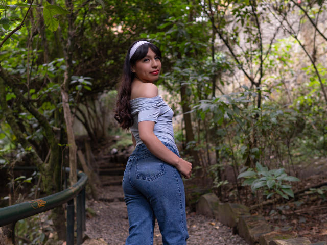 ZoeRedds with long dark hair, off-shoulder gray top, blue jeans, and white headband, standing on forest stairs, looking back, showing her ass, in a green, wooded area.