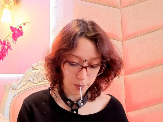 JinXkm with wavy brown hair, glasses, black choker, and black shirt, sucking on a straw, has a small cross on her forehead, and pinkish glow.