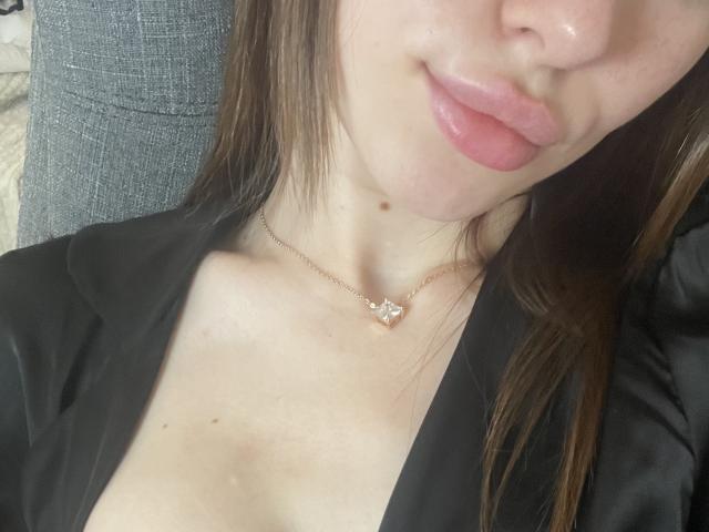 Il viso di Ramilla, con le labbra rosate e lucide, una piccola lentiggine sul collo e una delicata collana con una piccola gemma. Indossa una maglietta nera che lascia intravedere leggermente il seno..