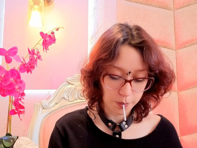 JinXkm with red curly hair, glasses, black choker, and a cigarette, has a cross tattoo on her forehead, wearing a black top, and pink orchids in the background.
