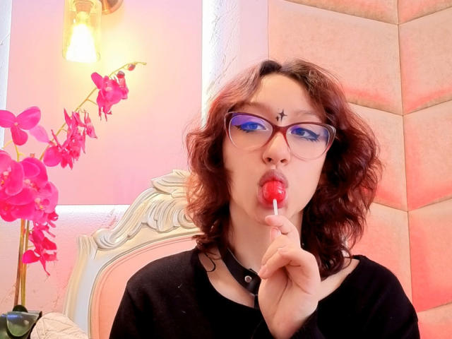 JinXkm with curly brown hair, blue eyeshadow, black choker, and glasses, sucking on a red lollipop, has a black star tattoo on her forehead, wearing a black top.