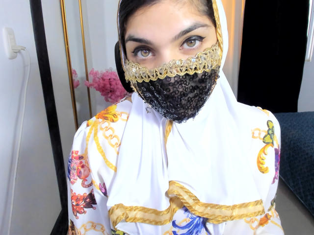 AniisaAli with brown eyes, wearing a white floral robe, gold trim, black lace face mask, and headscarf, looking at the camera, in a brightly lit room.