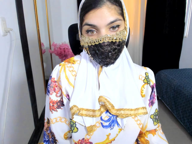 AniisaAli with dark hair, brown eyes, wearing a white floral hijab, golden lace mask, and white floral robe with gold and blue embroidery, sitting in a chair.