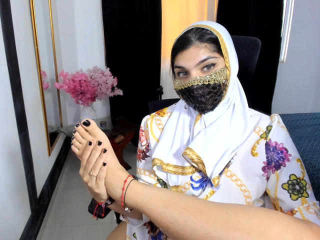 AniisaAli with dark makeup, wearing a white floral hijab, gold lace mask, and black nail polish, holding her foot with painted toenails. Her feet are in the foreground.