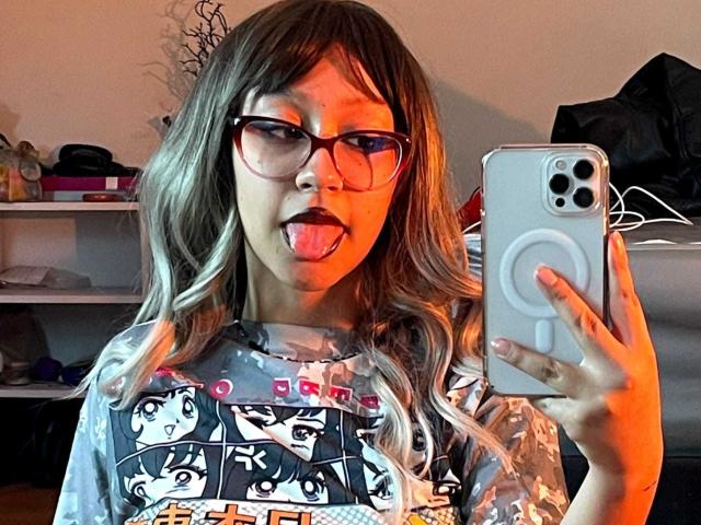 JinXkm with long blonde hair, glasses, and tongue out, holding a phone in a mirror selfie. Wearing a gray graphic tee, showing her cute face and small tits.