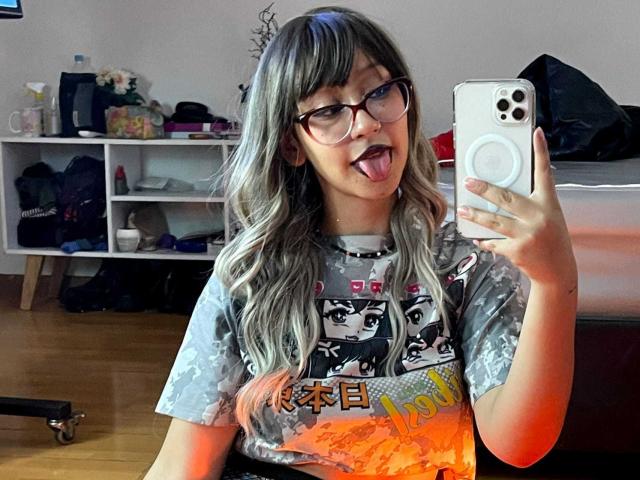 JinXkm with glasses, tongue out, holding phone, wearing gray graphic tee, long blonde and black hair, taking selfie in room. Boobs not visible. Ass not visible. Pussy not visible.
