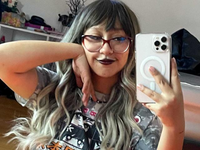 JinXkm with long, wavy gray hair, red glasses, and dark lipstick, holding a phone in front of her face, smiling, showing her tits in a mirror selfie.