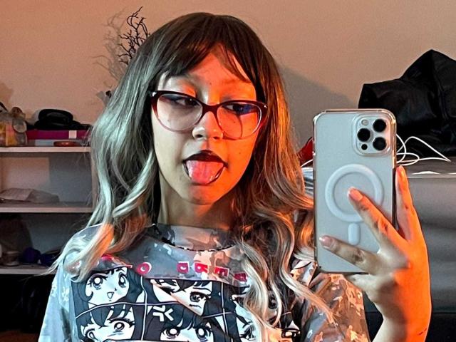 JinXkm with glasses, tongue out, holding a phone, wearing a graphic shirt, long wavy blonde hair, orange light, phone in right hand, mirror selfie.