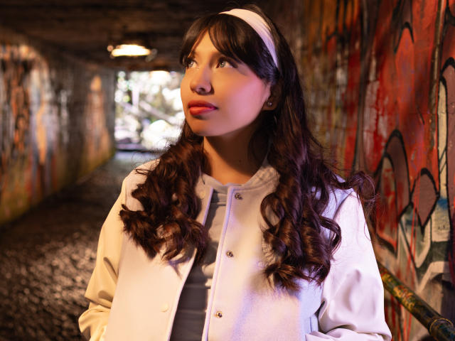 ZoeRedds a les cheveux bruns bouclés, un bandeau blanc, et porte un sweat à capuche blanc avec une fermeture boutonnée sur le devant. Elle se tient dans un tunnel faiblement éclairé, recouvert de graffitis, et regarde sur le côté..