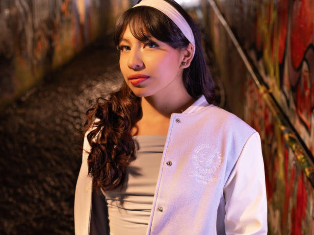ZoeRedds with long, wavy brown hair, wearing a white headband, buttoned-up white jacket, and grey dress, standing in a graffiti-covered, dimly-lit alley. Her face is slightly turned.