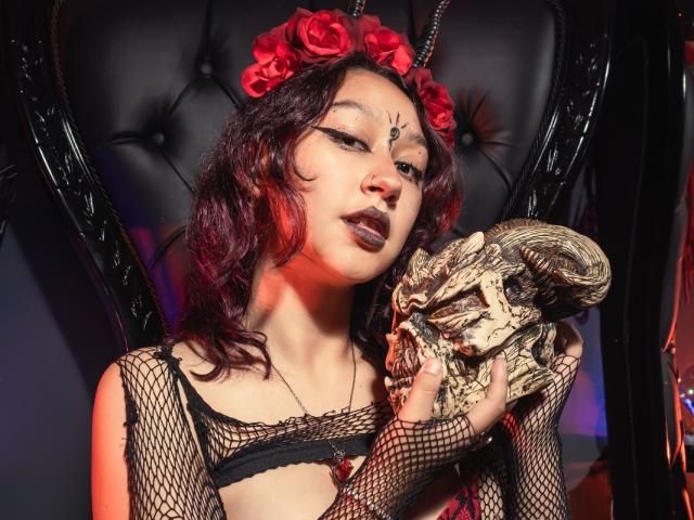 JinXkm with dark red lipstick and wavy hair, wearing a red flower crown and black fishnet top, holding a skull, sitting in a black, leather-upholstered chair.