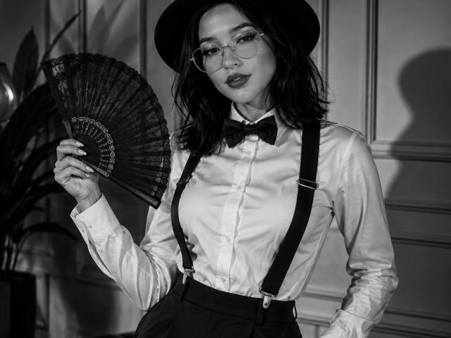 Celeste Ocampo con occhiali, cappello nero, camicia bianca, papillon nero, bretelle, che tiene un ventaglio scuro, mostra il sedere, seno di media grandezza, capelli scuri, posa sicura, foto in bianco e nero..