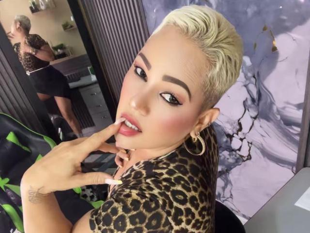 Gianella Cox, con cabello rubio corto, vistiendo una blusa de estampado de leopardo, tocándose los labios rosados con un dedo, mirando a la cámara, mostrando sus tatuajes y aros de oro..