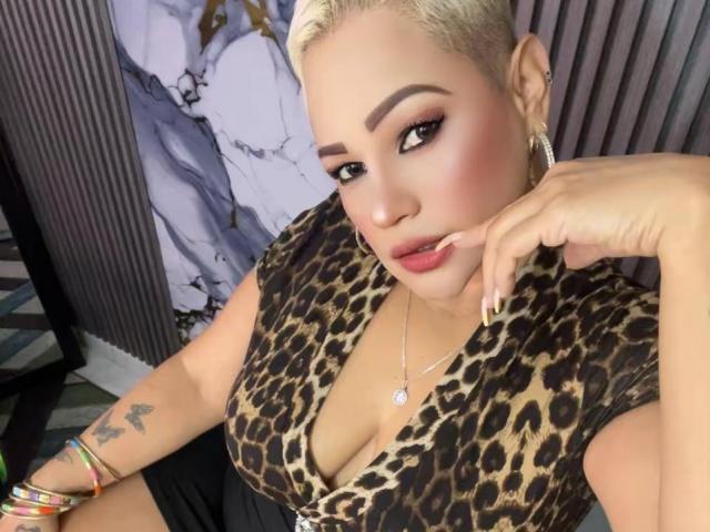 Gianella Cox, con cabello rubio, maquillaje llamativo y aros grandes, vestida con una blusa de estampado de leopardo, mostrando sus senos, y brazaletes coloridos, tocándose los labios..