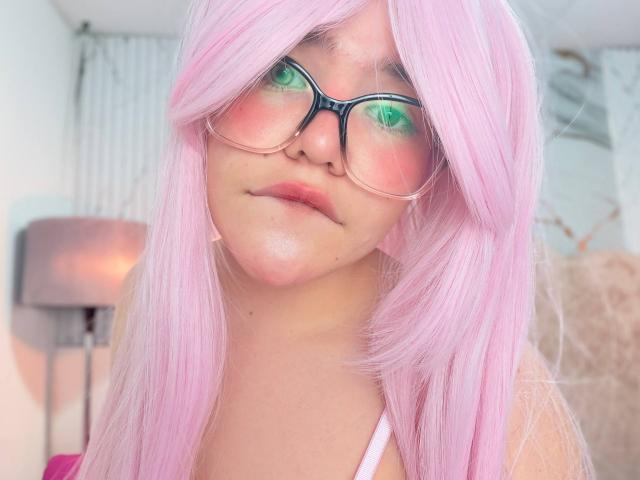 AlexaMissFortune con cabello rosa, gafas verdes y rubor rosa, mirando a la cámara con un ligero ceño fruncido, mostrando sus pechos con una blusa blanca..