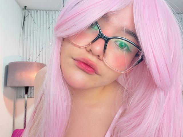 AlexaMissFortune with pink hair, green eyeshadow, and glasses, has pink lips and a slightly pouty expression. Her tits are not visible. She's in a soft-lit indoor setting.