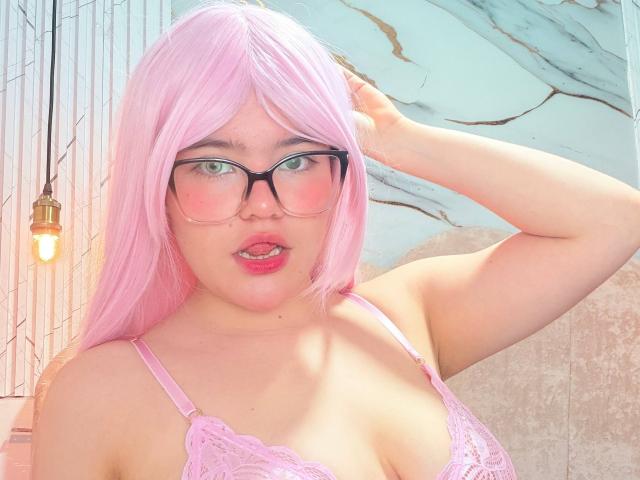 AlexaMissFortune with pink hair, glasses, and pink lace bra, showing her tits, has a hand in her hair, pink lips, and a light bulb in the background.