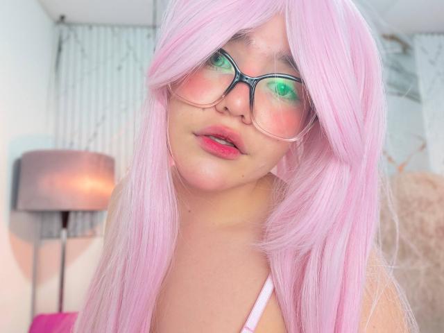 AlexaMissFortune with pink hair, green-tinted glasses, and pink lipstick, showing a slightly open mouth. Her tits are not visible, but she's wearing a white top.