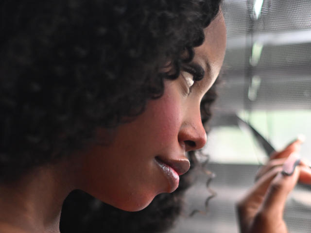 CatalinaSanA with curly black hair, dark brown skin, and a soft smile, looking to the side, holding a pen near her face, in a semi-soft lit profile.